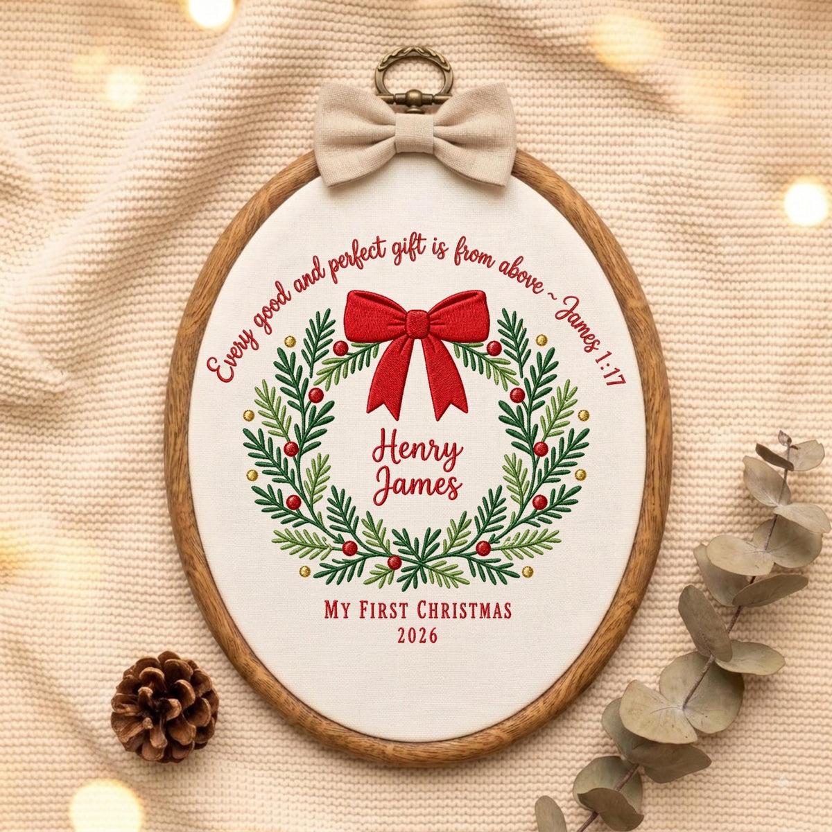May include: An oval wooden embroidery hoop containing white fabric with a red and green embroidered wreath, a red bow, and the text: Every good and perfect gift is from above ~ James 1:17, Henry James, MY FIRST CHRISTMAS, 2026. A beige bow is attached.