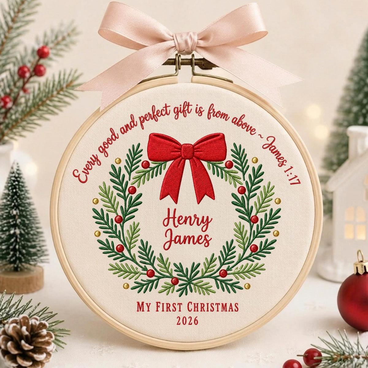 May include: A circular wooden embroidery hoop containing cream fabric with a stitched Christmas wreath, a red bow, and the text "Every good and perfect gift is from above ~ James 1:17", "Henry James", "MY FIRST CHRISTMAS", and "2026". A pale pink ribbon is tied at the top.