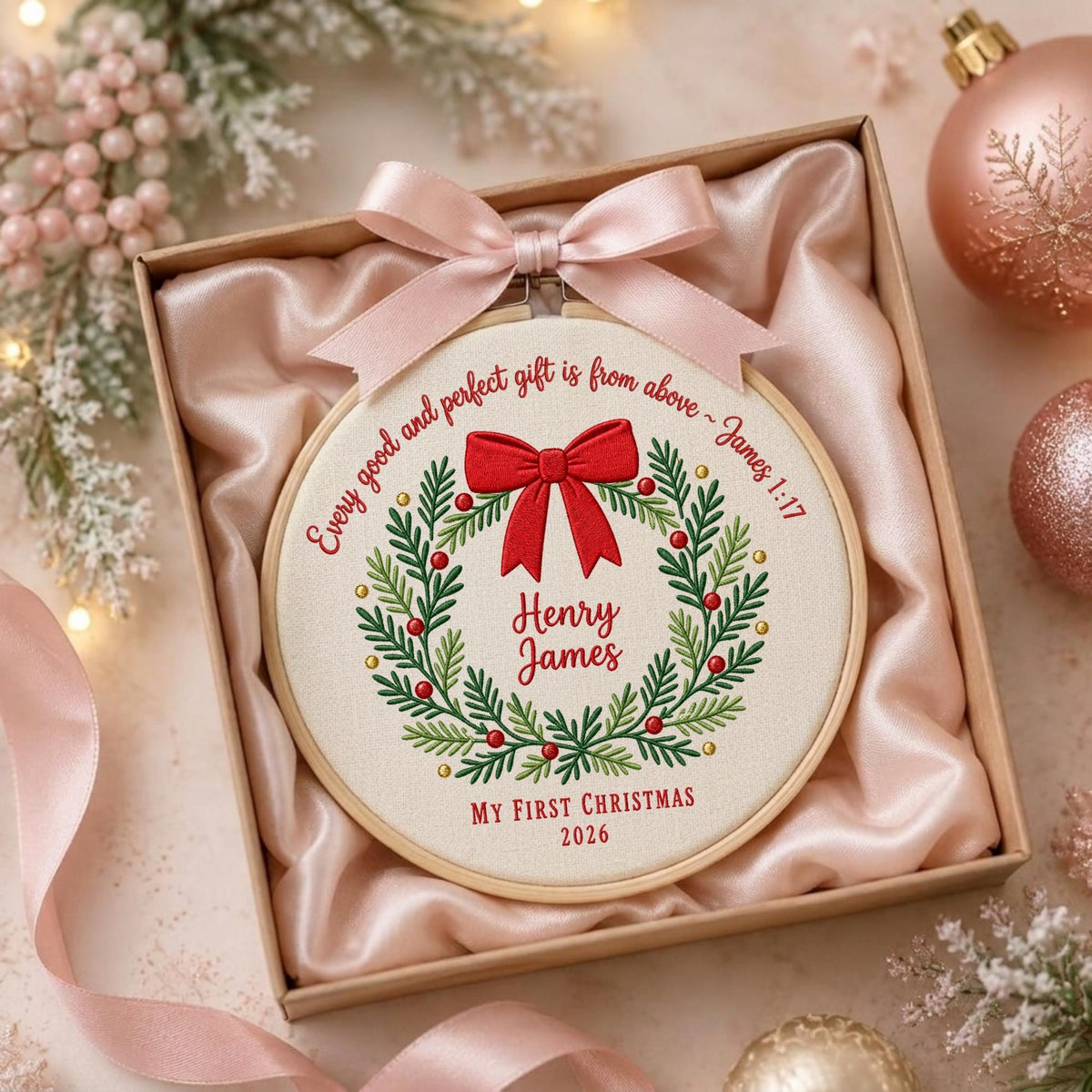 May include: A circular embroidery hoop containing white fabric with a stitched green wreath, a red bow, and red berries. The text reads: Every good and perfect gift is from above ~ James 1:17, Henry James, and MY FIRST CHRISTMAS 2026. A pink ribbon is tied to the top.