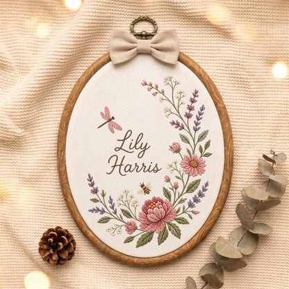 May include: An oval wooden embroidery hoop containing white fabric with the embroidered name Lily Harris, a pink dragonfly, a small bee, and a floral wreath of pink flowers, purple lavender, and green leaves, topped with a beige fabric bow.
