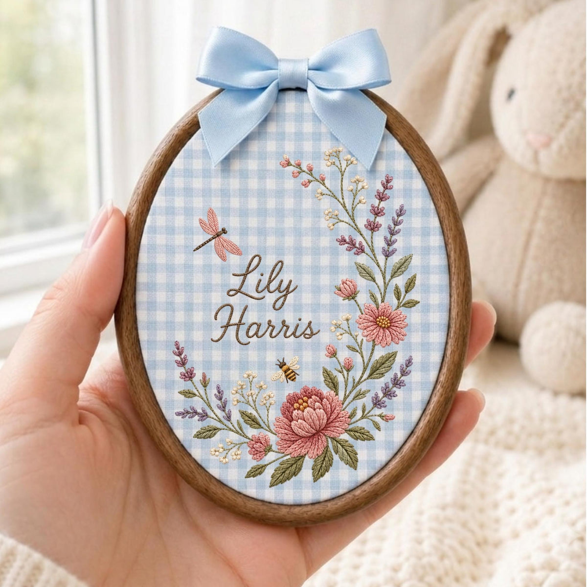 May include: An oval embroidery hoop with a dark wood frame, featuring light blue and white gingham fabric. The center displays the name Lily Harris in brown script, surrounded by embroidered pink flowers, lavender, a dragonfly, and a bee, topped with a light blue satin bow.