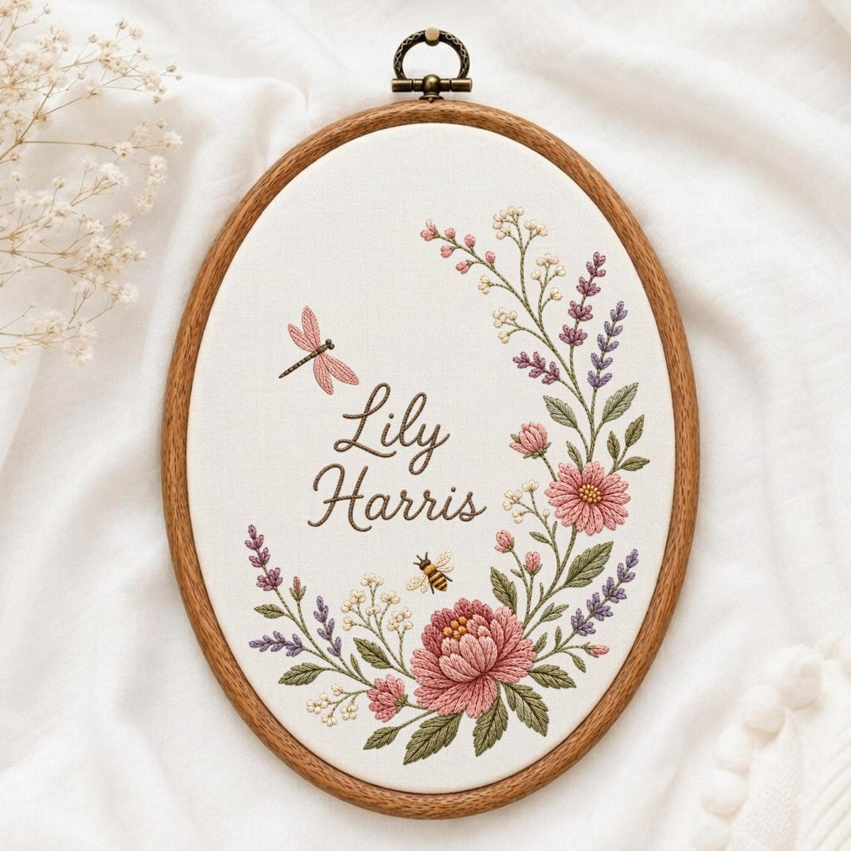 May include: An oval wooden embroidery hoop containing white fabric with the embroidered name Lily Harris. The design features a floral wreath with pink and purple flowers, green leaves, a small bee, and a pink dragonfly in a cursive font.