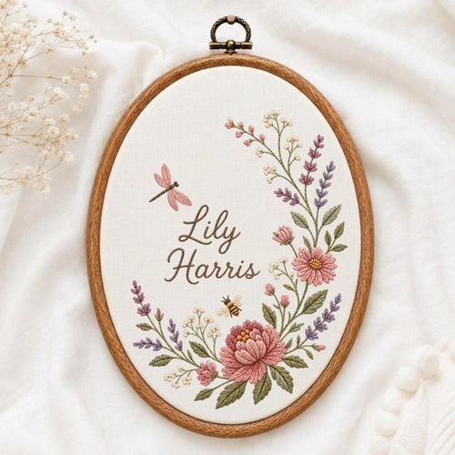 May include: An oval wooden embroidery hoop containing white fabric with the embroidered name Lily Harris. The design features a floral wreath with pink and purple flowers, green leaves, a small bee, and a pink dragonfly in a cursive font.