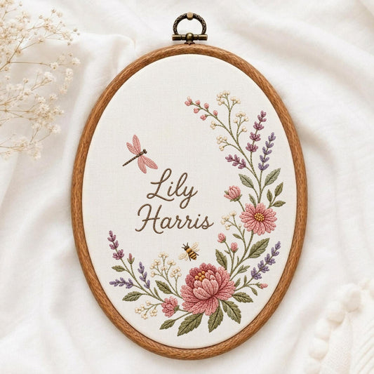 May include: An oval wooden embroidery hoop containing white fabric with the embroidered name Lily Harris. The design features a floral wreath with pink and purple flowers, green leaves, a small bee, and a pink dragonfly in a cursive font.