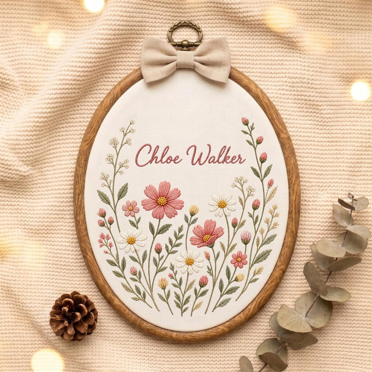 May include: An oval wooden embroidery hoop containing white fabric with hand-stitched floral designs in pink, white, and yellow. The name Chloe Walker is embroidered in pink script above the flowers. A beige fabric bow is attached to the top.