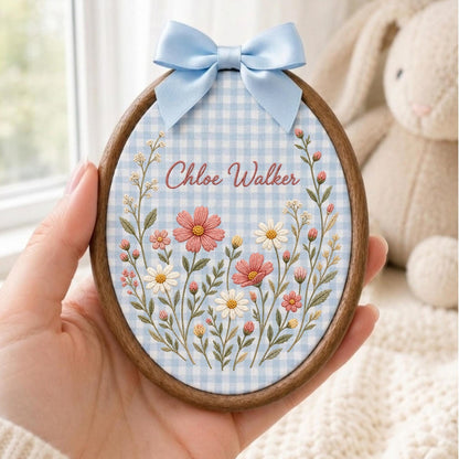 May include: An oval embroidery hoop featuring a light blue and white gingham fabric background. The fabric is embroidered with a floral arrangement of pink and white flowers and green stems. The name Chloe Walker is stitched in pink thread.