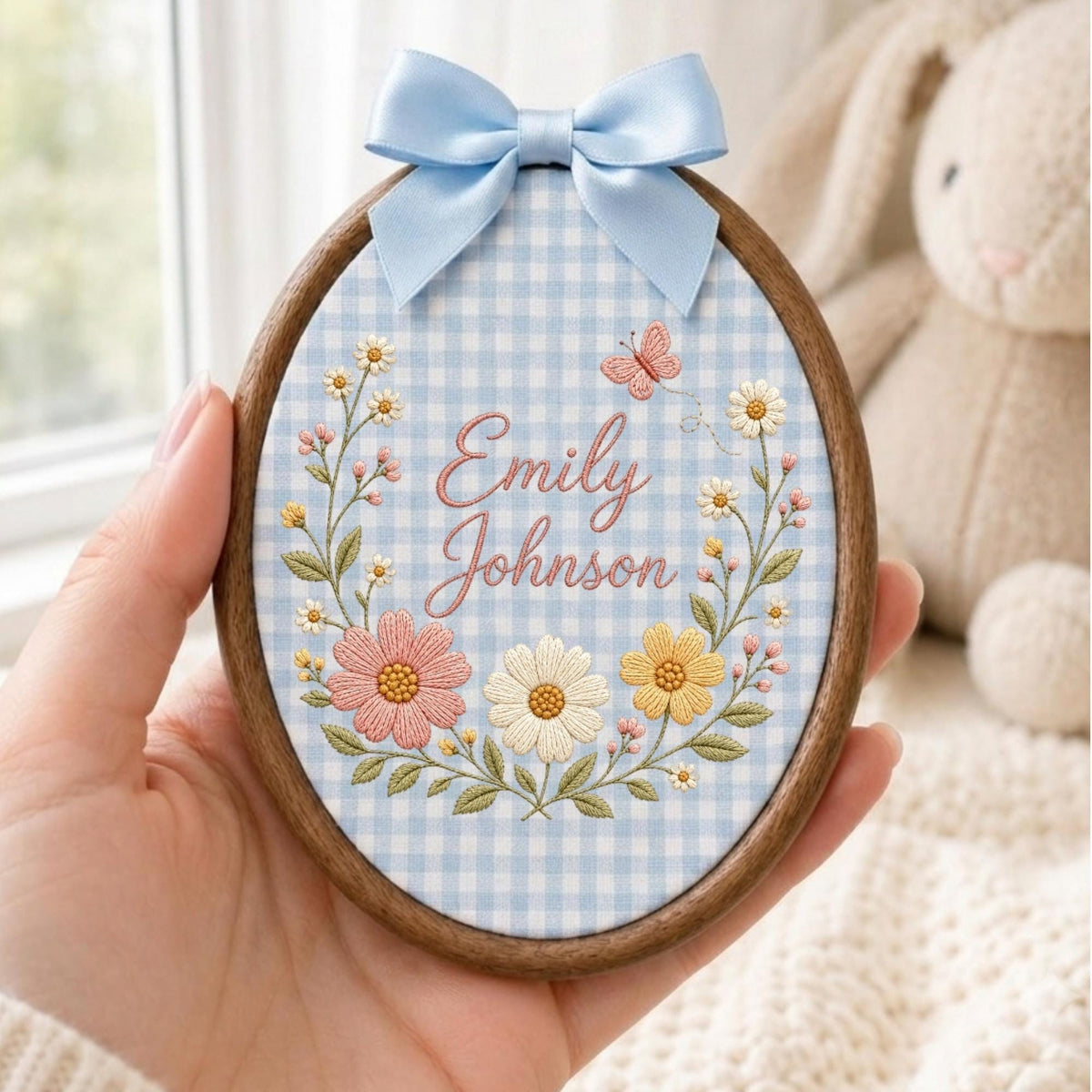 May include: An oval wooden embroidery hoop containing light blue and white gingham fabric. The fabric features embroidered flowers in pink, white, and yellow, a small butterfly, and the name Emily Johnson in pink script. A light blue satin bow is attached.