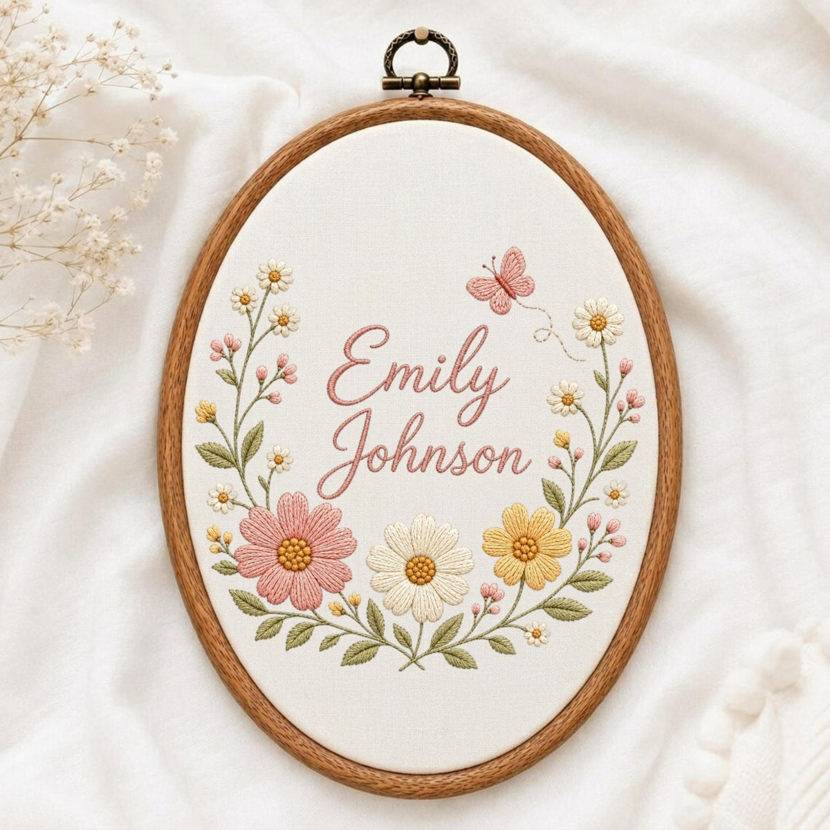 May include: An oval wooden embroidery hoop containing white fabric with the embroidered name Emily Johnson in pink cursive script. The design features a floral wreath of pink, yellow, and white flowers with green leaves and a small pink butterfly.