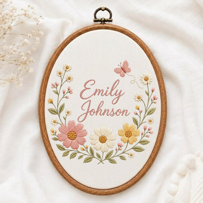 May include: An oval wooden embroidery hoop containing white fabric with the embroidered name Emily Johnson in pink cursive script. The design features a floral wreath of pink, yellow, and white flowers with green leaves and a small pink butterfly.