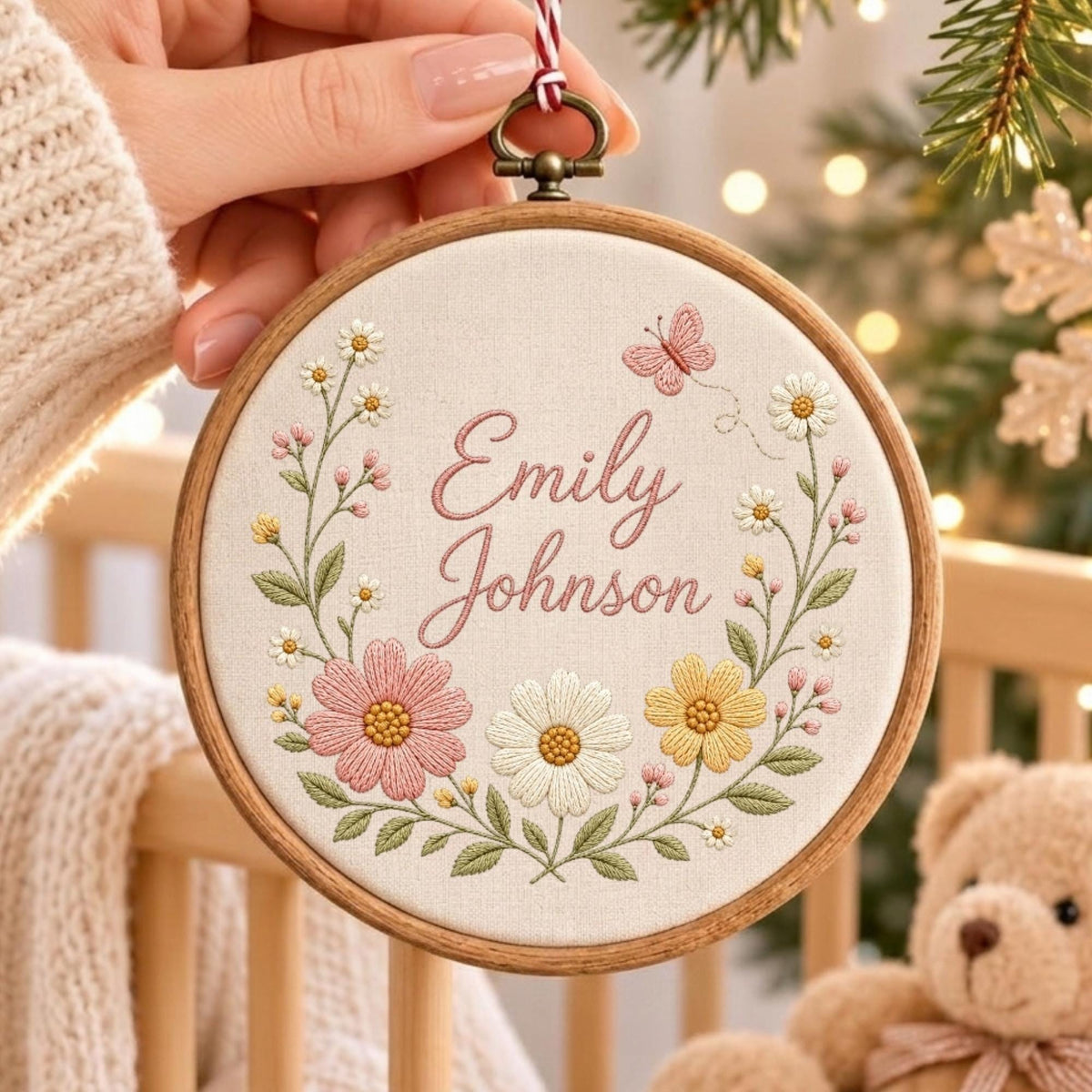 May include: A circular wooden embroidery hoop containing off-white fabric with the embroidered name Emily Johnson in pink script. The design features a floral wreath with pink, white, and yellow flowers, green leaves, and a small pink butterfly.