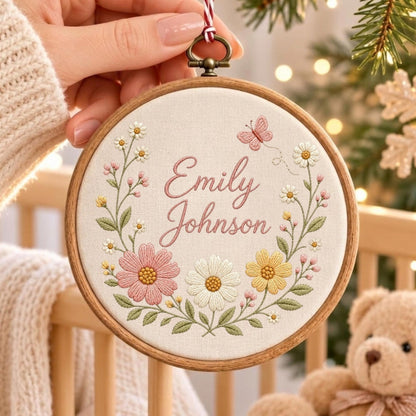 May include: A circular wooden embroidery hoop containing off-white fabric with the embroidered name Emily Johnson in pink script. The design features a floral wreath with pink, white, and yellow flowers, green leaves, and a small pink butterfly.