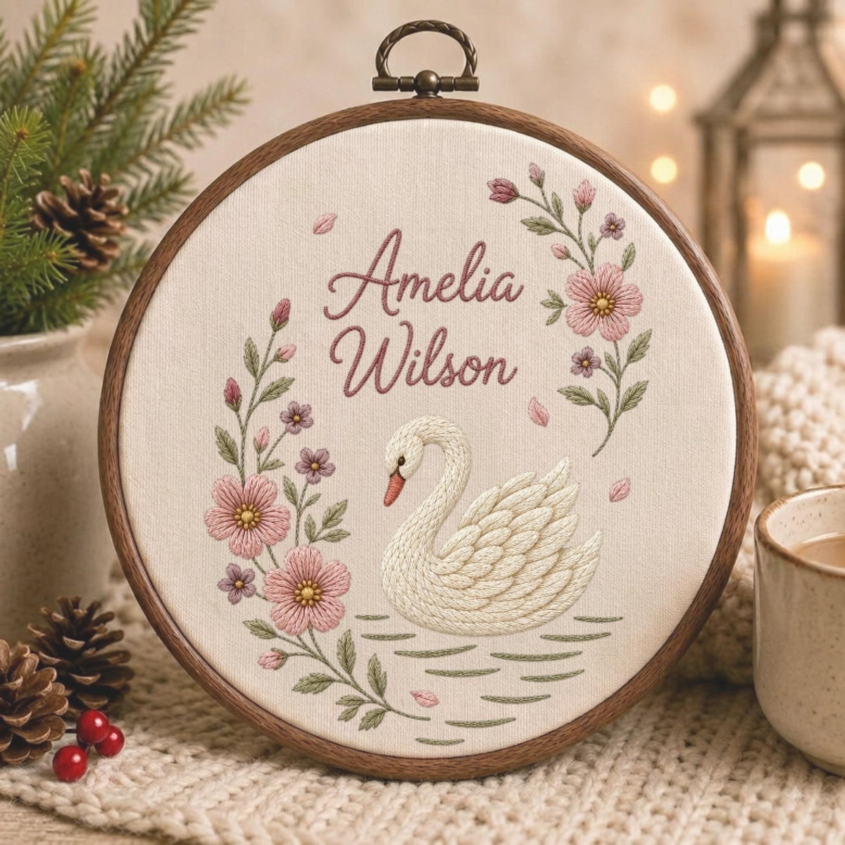 May include: A circular wooden embroidery hoop containing cream-colored fabric with a hand-stitched design. The embroidery features a white swan swimming on water, surrounded by pink flowers and green leaves, with the name Amelia Wilson in script.