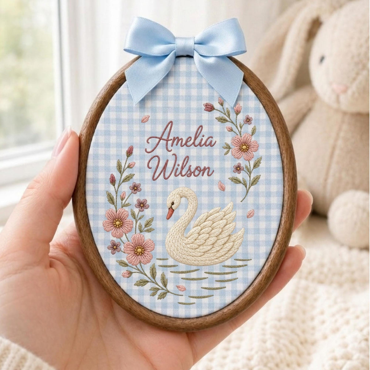 May include: An oval wooden embroidery hoop containing fabric with a light blue and white gingham pattern. The fabric features an embroidered white swan, pink flowers, and the text Amelia Wilson in brown script, topped with a light blue satin bow.