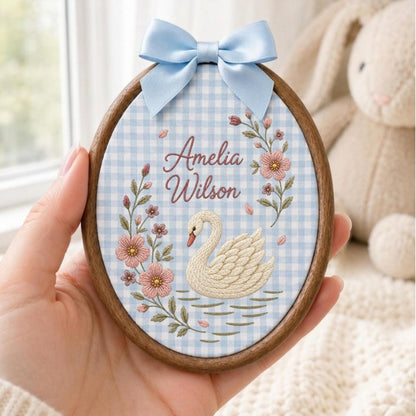May include: An oval wooden embroidery hoop containing fabric with a light blue and white gingham pattern. The fabric features an embroidered white swan, pink flowers, and the text Amelia Wilson in brown script, topped with a light blue satin bow.