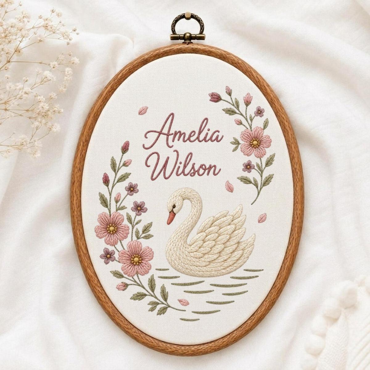 May include: An oval wooden embroidery hoop containing white fabric with a hand-stitched design of a white swan on water, pink flowers, and the text Amelia Wilson in dark pink script. A metal hanging loop is attached to the top of the frame.