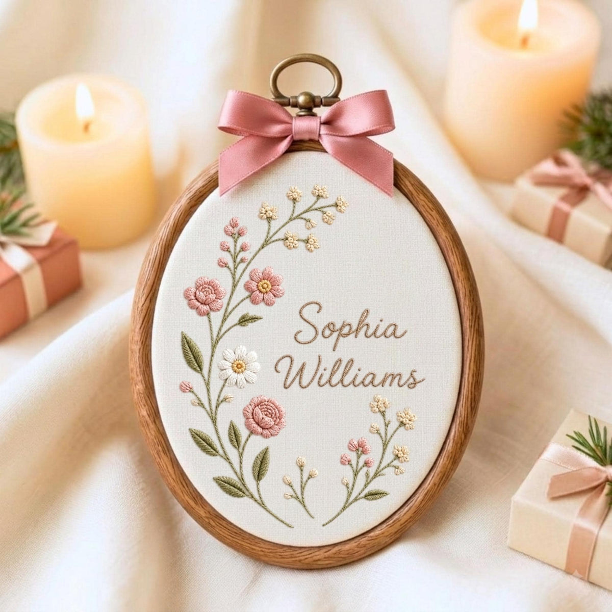May include: An oval wooden embroidery hoop containing white fabric with the embroidered name Sophia Williams in brown script. A floral arrangement of pink and white flowers with green leaves curves along the left side. A dusty pink satin bow is attached.