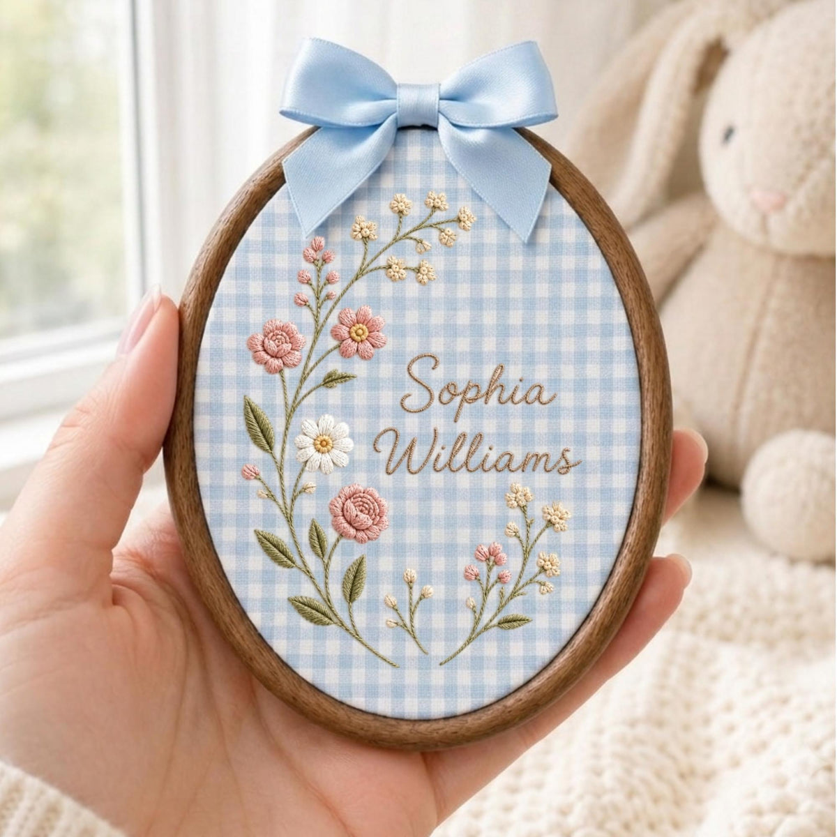May include: An oval wooden embroidery hoop containing light blue and white gingham fabric. The fabric features embroidered pink and white flowers with green stems and the text Sophia Williams in brown script. A light blue satin bow sits on top.