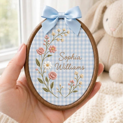 May include: An oval wooden embroidery hoop containing light blue and white gingham fabric. The fabric features embroidered pink and white flowers with green stems and the text Sophia Williams in brown script. A light blue satin bow sits on top.