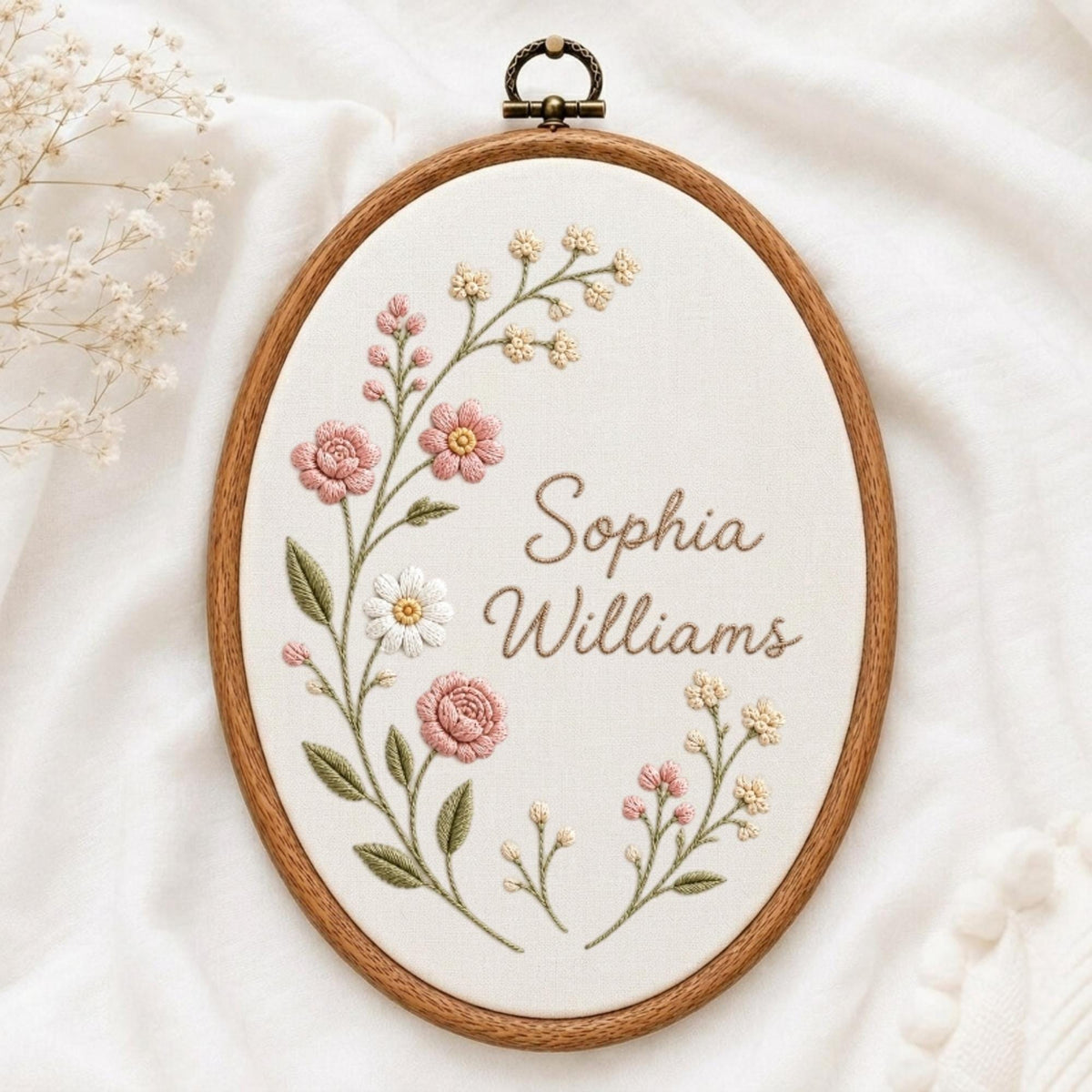 May include: An oval wooden embroidery hoop containing white fabric with hand-stitched floral designs in pink, white, and green. The name Sophia Williams is embroidered in brown script in the center. A metal hanging loop is attached to the top.