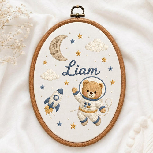 May include: An oval embroidery hoop containing white fabric with a space-themed design. The embroidery features a brown bear in a white astronaut suit, a blue and white rocket, a crescent moon, clouds, and yellow and blue stars with the name Liam.