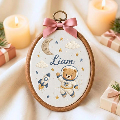May include: An oval wooden embroidery hoop featuring a white fabric background with a hand-stitched design of a teddy bear astronaut, a rocket ship, a crescent moon, clouds, and stars. The name Liam is embroidered in blue script, topped with a pink bow.