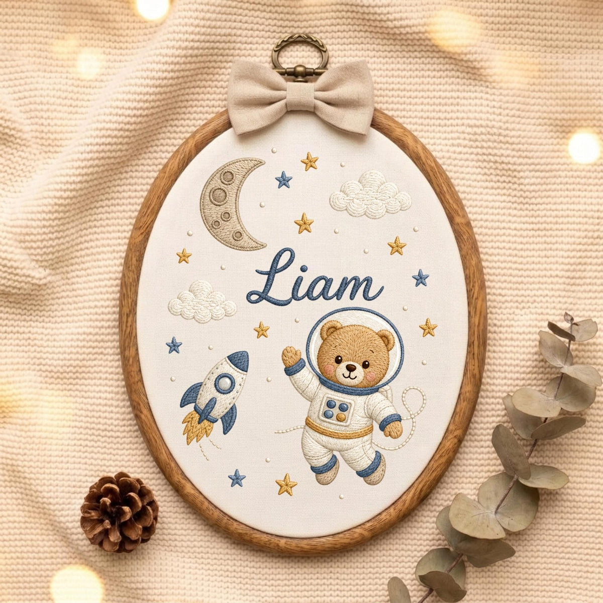 May include: An oval embroidery hoop featuring a white fabric background with a brown teddy bear astronaut, a blue and white rocket, a crescent moon, clouds, and yellow and blue stars. The name Liam is embroidered in blue script in the center.