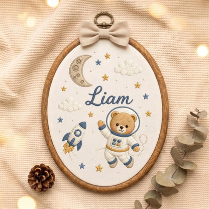 May include: An oval embroidery hoop featuring a white fabric background with a brown teddy bear astronaut, a blue and white rocket, a crescent moon, clouds, and yellow and blue stars. The name Liam is embroidered in blue script in the center.