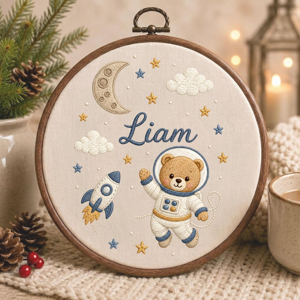 May include: A circular wooden embroidery hoop containing a fabric panel with a space-themed design. The embroidery features a brown teddy bear in a white astronaut suit, a blue and white rocket, a crescent moon, clouds, and stars with the name Liam.