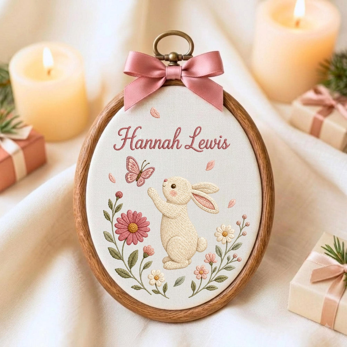 May include: An oval wooden embroidery hoop containing white fabric with the embroidered name Hannah Lewis, a cream-colored rabbit, a pink butterfly, and various pink and white flowers. A dusty pink satin ribbon bow is attached to the top.