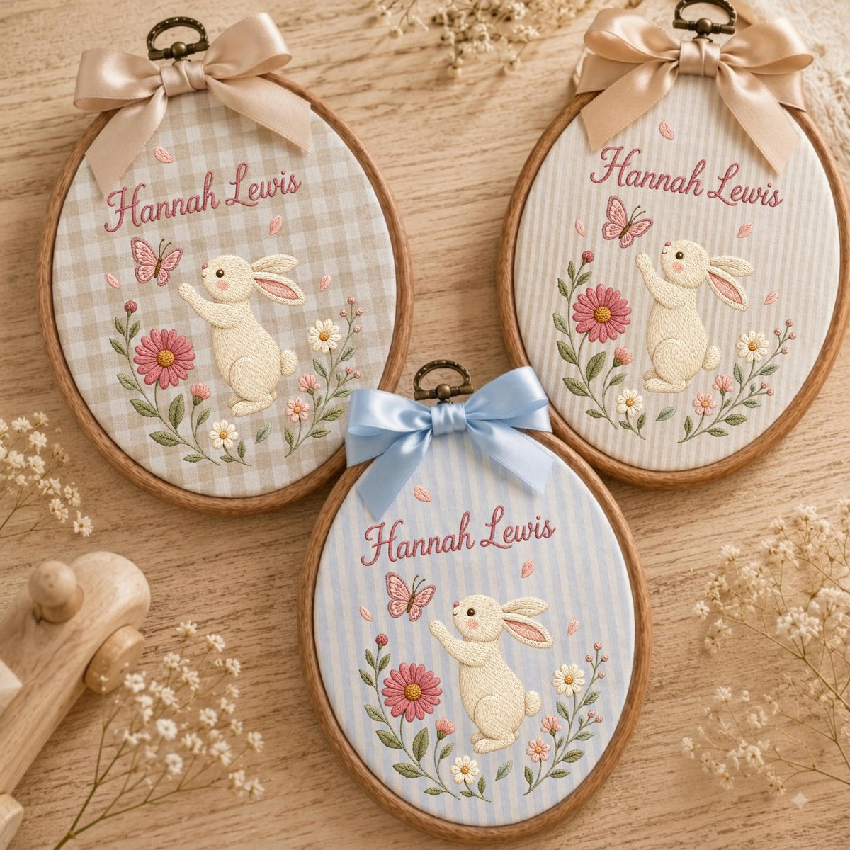 May include: Three oval wooden embroidery hoops featuring fabric backgrounds with gingham, striped, and pinstriped patterns. Each hoop displays embroidered cream-colored rabbits, pink flowers, butterflies, and the name Hannah Lewis in script.