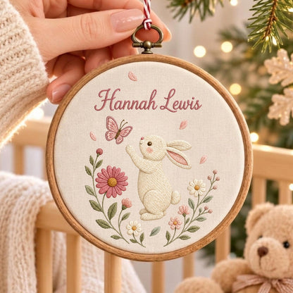May include: A circular wooden embroidery hoop containing a cream-colored fabric panel. The fabric features an embroidered design of a white rabbit reaching toward a pink butterfly, surrounded by pink and white flowers, with the name Hannah Lewis.