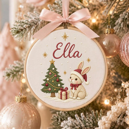 May include: A circular embroidery hoop ornament featuring the name Ella in red script. The design includes a green Christmas tree with ornaments, a gift box, and a beige rabbit wearing a red Santa hat and bow, topped with a pale pink ribbon.