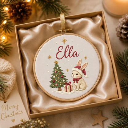 May include: A round embroidery hoop ornament featuring a cream fabric background with the name Ella embroidered in red. The design includes a green Christmas tree with colorful ornaments, a gift box, and a rabbit wearing a red winter hat.