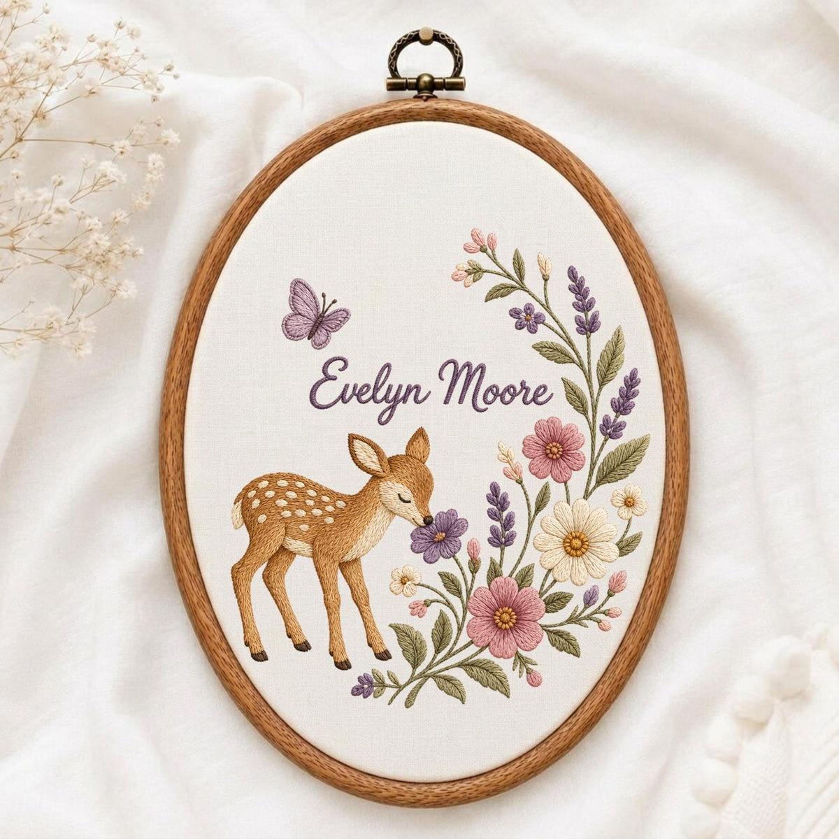 May include: An oval wooden embroidery hoop containing white fabric with a hand-stitched design of a brown fawn, a purple butterfly, and a floral arrangement of pink, white, and purple flowers. The text Evelyn Moore is stitched in purple.