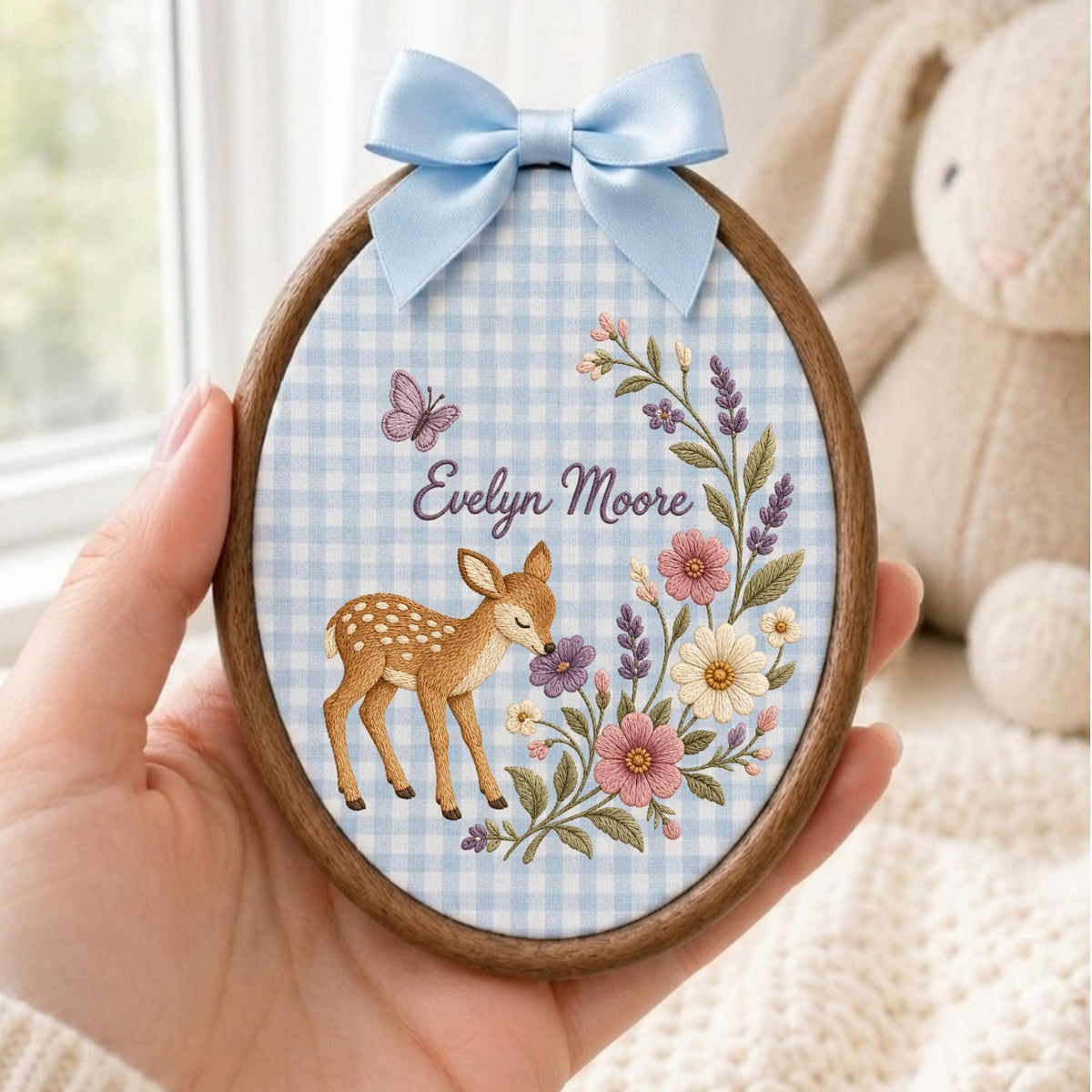 May include: An oval wooden embroidery hoop featuring a light blue and white gingham fabric background. The fabric is embroidered with a brown fawn, a purple butterfly, and a floral spray. The name Evelyn Moore is stitched in purple script.