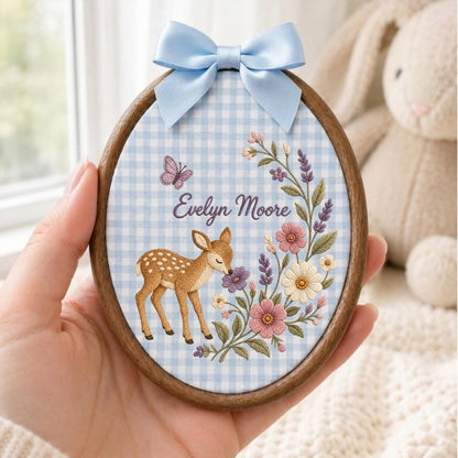 May include: An oval wooden embroidery hoop featuring a light blue and white gingham fabric background. The fabric is embroidered with a brown fawn, a purple butterfly, and a floral spray. The name Evelyn Moore is stitched in purple script.