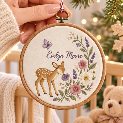 May include: A round wooden embroidery hoop containing off-white fabric with a hand-stitched design of a brown fawn, a purple butterfly, and a floral arrangement in shades of pink, white, and purple. The text Evelyn Moore is stitched in purple.