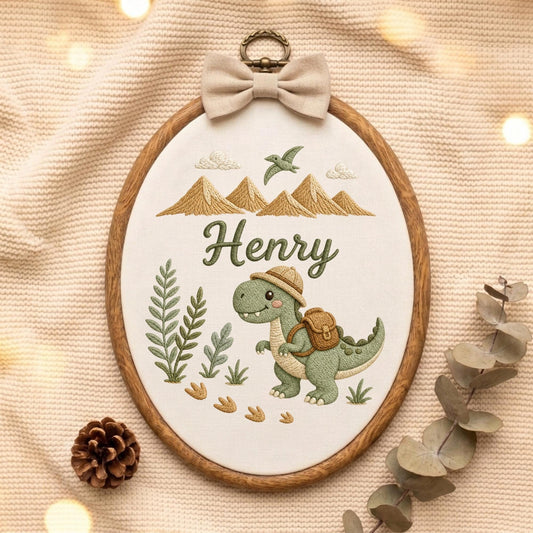 May include: An oval embroidery hoop containing white fabric with a stitched design of a green dinosaur wearing a hat and backpack, the name Henry, mountains, and plants. A beige fabric bow is attached to the top of the wooden frame.