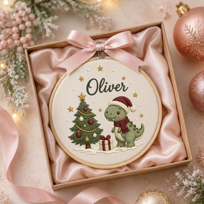 May include: A round wooden embroidery hoop containing fabric with an embroidered green dinosaur wearing a red Santa hat and scarf, a decorated Christmas tree, a gift box, and the name Oliver in dark green script, topped with a pink satin bow.