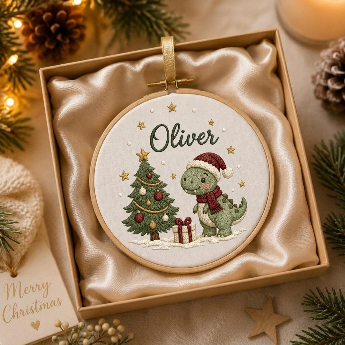 May include: A round wooden embroidery hoop containing white fabric with a stitched design of a green dinosaur wearing a red scarf and Santa hat next to a decorated Christmas tree and a gift, with the name Oliver embroidered in dark green text.