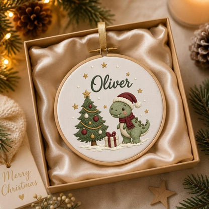 May include: A round wooden embroidery hoop containing white fabric with a stitched design of a green dinosaur wearing a red scarf and Santa hat next to a decorated Christmas tree and a gift, with the name Oliver embroidered in dark green text.