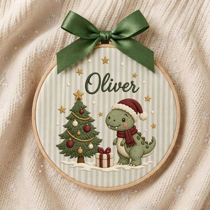 May include: A round wooden embroidery hoop featuring a light green and white striped fabric background. The design includes the name Oliver, a decorated Christmas tree, a gift box, and a green dinosaur wearing a Santa hat and scarf, topped with a green bow.