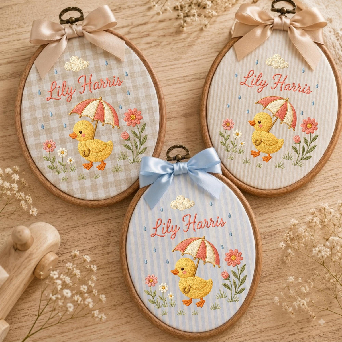 May include: Three oval wooden embroidery hoops featuring fabric with embroidered yellow ducks holding umbrellas, pink flowers, and the name Lily Harris. Each hoop is topped with a satin ribbon bow in beige or light blue, displaying a charming design.