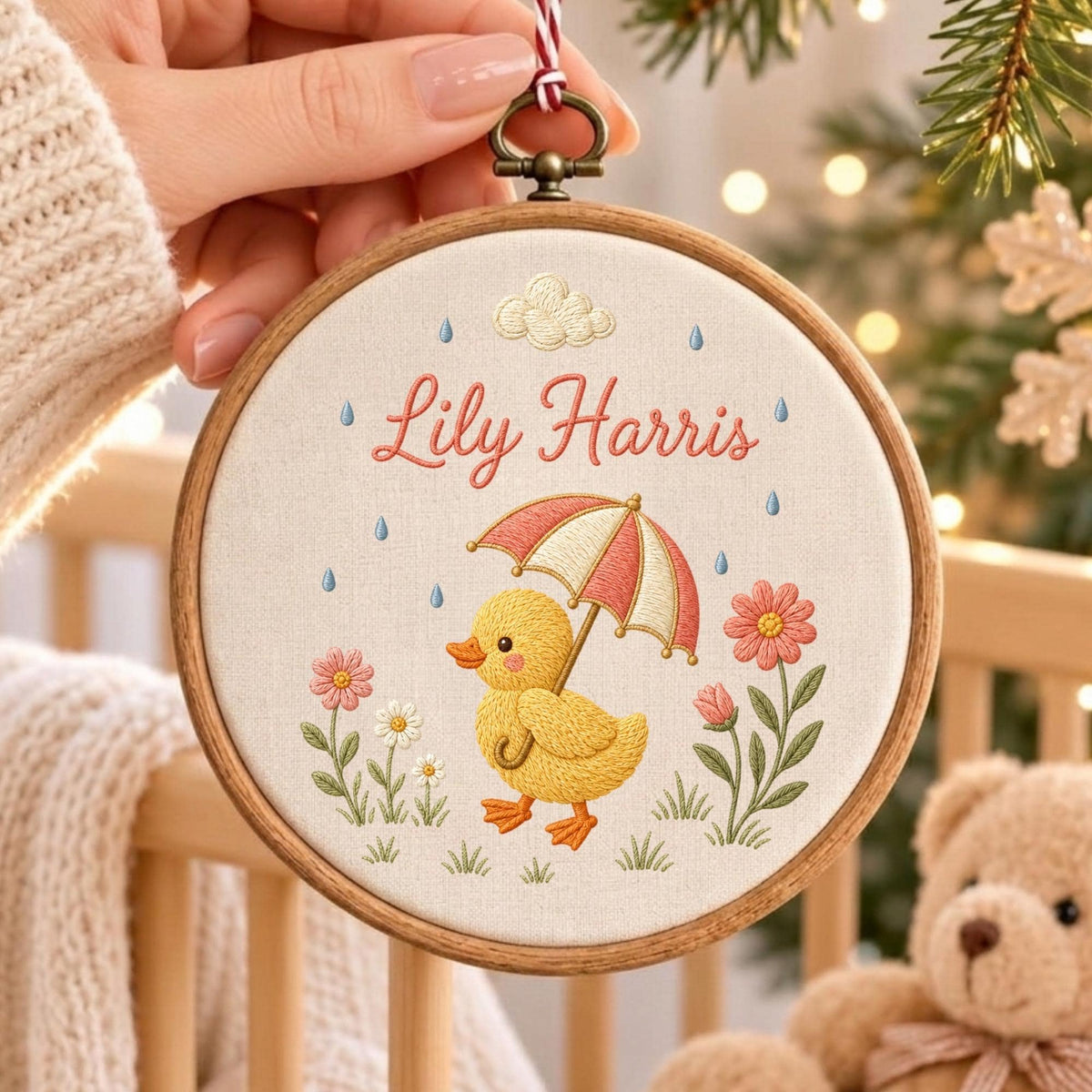 May include: A circular wooden embroidery hoop containing a cream-colored fabric panel. The embroidery features a yellow duckling holding a red and white striped umbrella, surrounded by pink and white flowers, blue raindrops, and a cloud, with the name Lily Harris stitched in red.