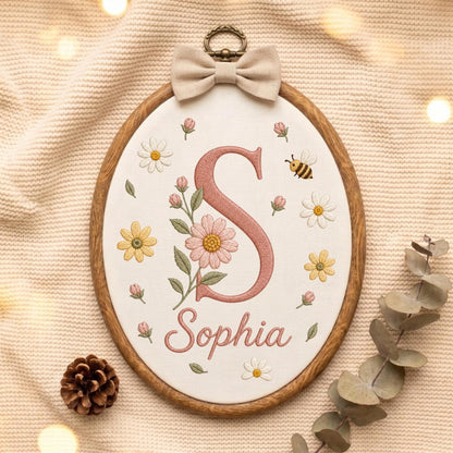 May include: An oval wooden embroidery hoop containing white fabric with a large embroidered letter S in dusty pink, the name Sophia in cursive, and floral motifs including daisies and a small bee. A beige fabric bow is attached to the top.