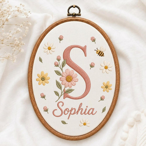 May include: An oval embroidery hoop containing white fabric with a large, dusty pink embroidered letter S, the name Sophia in matching pink script, and surrounding floral motifs in yellow and pink with green leaves and a small bumblebee.