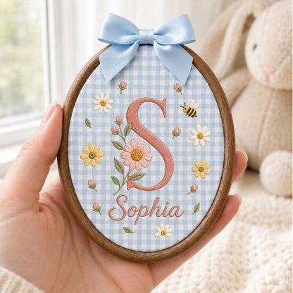 May include: An oval wooden embroidery hoop featuring light blue gingham fabric. The design includes a large embroidered letter S in dusty rose, the name Sophia in cursive, and floral motifs with daisies, buds, and a small bee, topped with a blue bow.