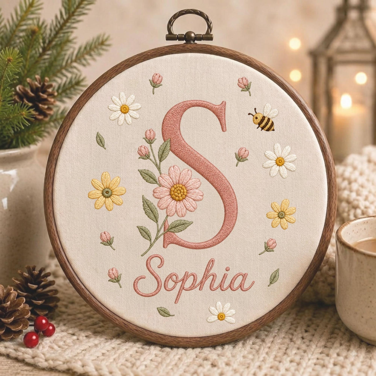 May include: A circular wooden embroidery hoop containing off-white fabric with a large pink letter S, the name Sophia in pink cursive script, and embroidered floral motifs including daisies, pink buds, green leaves, and a small bumblebee.