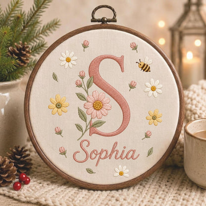 May include: A circular wooden embroidery hoop containing off-white fabric with a large pink letter S, the name Sophia in pink cursive script, and embroidered floral motifs including daisies, pink buds, green leaves, and a small bumblebee.