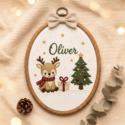 May include: An oval wooden embroidery hoop containing white fabric with a stitched design of a brown reindeer wearing a red scarf, a decorated green Christmas tree, a gift box, and gold stars. The name Oliver is embroidered in green script.